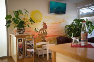 Start ein warteraum mit mehreren pflanzen, einem tisch mit zeitschriften und einem wandgemälde von simba aus könig der löwen, der bei sonnenaufgang auf dem königsfelsen steht. Über dem wandbild ist ein fernseher angebracht, und auf einem tisch im vordergrund steht eine wasserflasche.