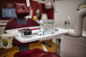 Start eine nahaufnahme eines zahnärztlichen stuhls zeigt ein tablett mit zahnärztlichem zubehör, darunter ein behälter mit watterollen, eine metallbox und pappbecher, in einer zahnarztpraxis mit kastanienbraunen wänden und medizinischen geräten.