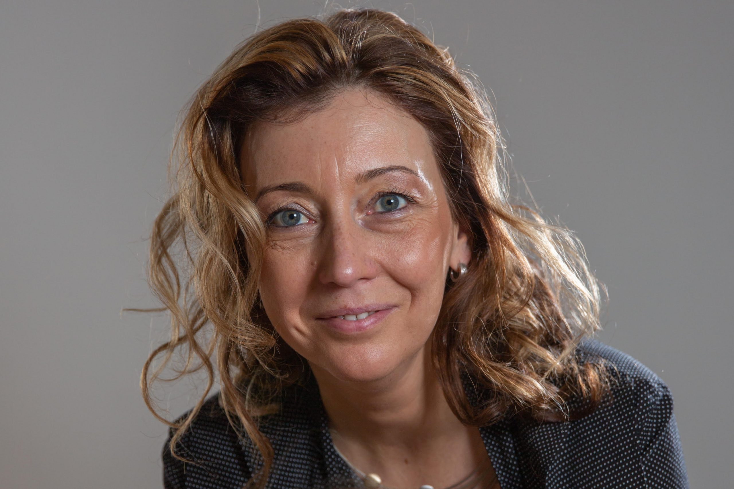 eine frau mit gewelltem hellbraunem haar, blauen augen und einem freundlichen lächeln trägt einen dunklen blazer und eine halskette und posiert vor einem schlichten grauen hintergrund.