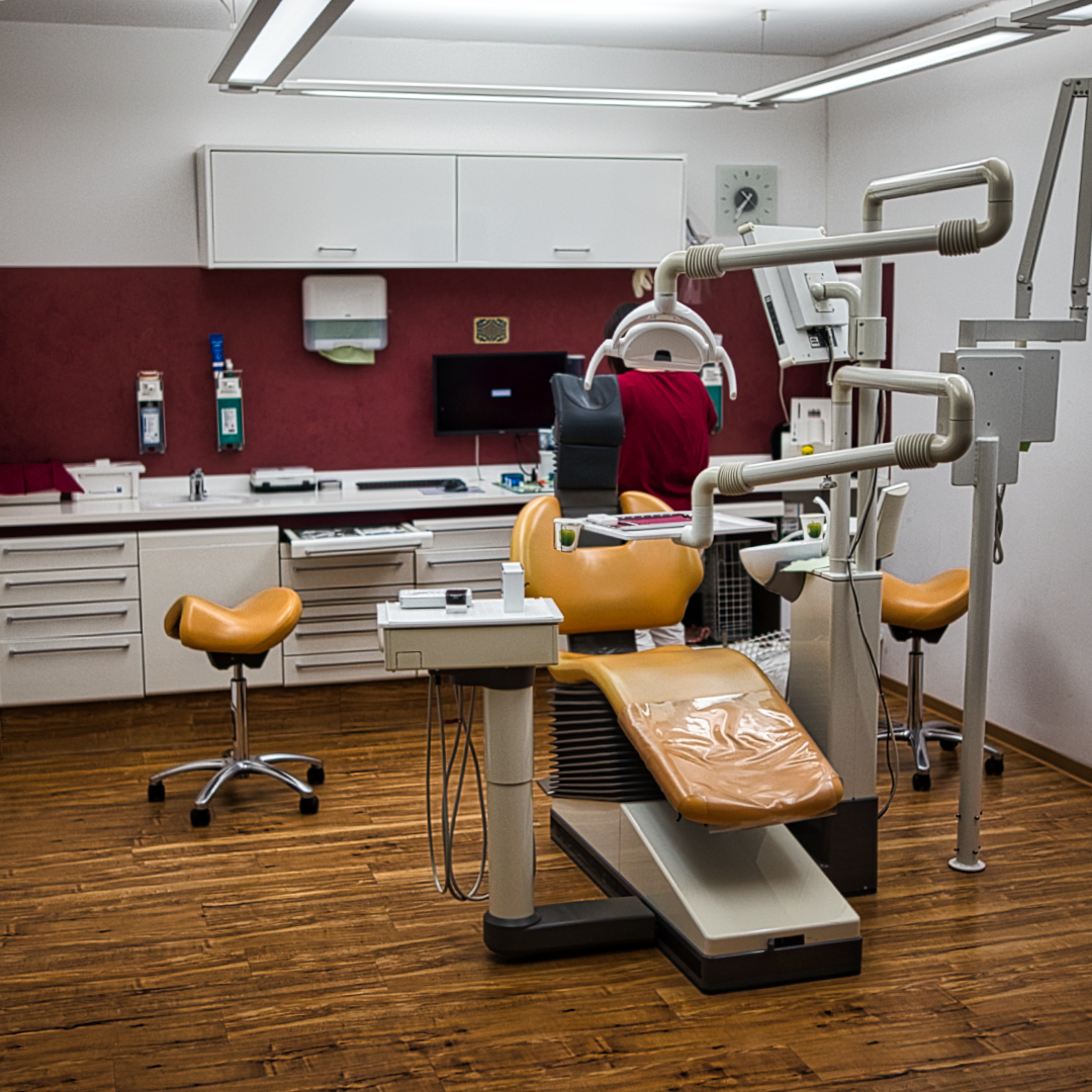 ein modernes zahnarztzimmer mit einem orangefarbenen zahnarztstuhl, oberlicht, computermonitor und zahnmedizinischen geräten. der raum hat holzböden, rote und weiße wände und schränke mit verschiedenen medizinischen geräten.